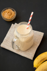 Mason glass jar mug filled with peanut butter banana smoothie on a black surface, low angle view. Close-up.