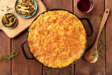 Homemade cottage pie in a cooking pan with pickles and wine, shot from above on a dark rustic wooden background