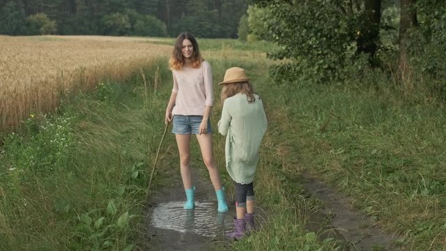Two happy girls sisters in boots playing in puddle of rainwater on country road, summer nature background after rain
