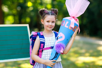 Happy little kid girl by desk with backpack or satchel and big school bag or cone traditional in...