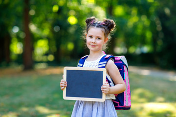 Happy little kid girl standing with desk and backpack or satchel. Schoolkid on first day of...