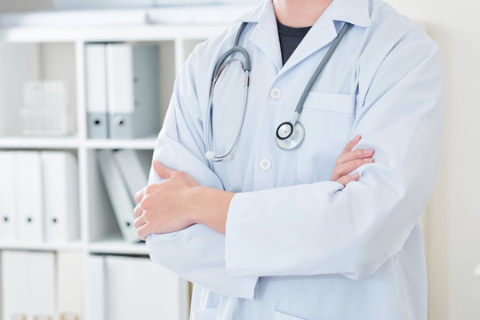 Cropped image of young doctor in white labcoat standing with his arms crossed in his office