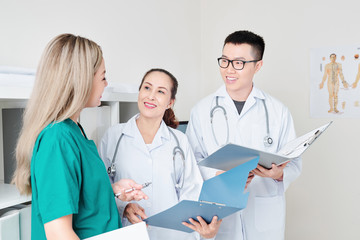 Smiling Vietnamese physicians standing in medical office with opened folders and discussing news