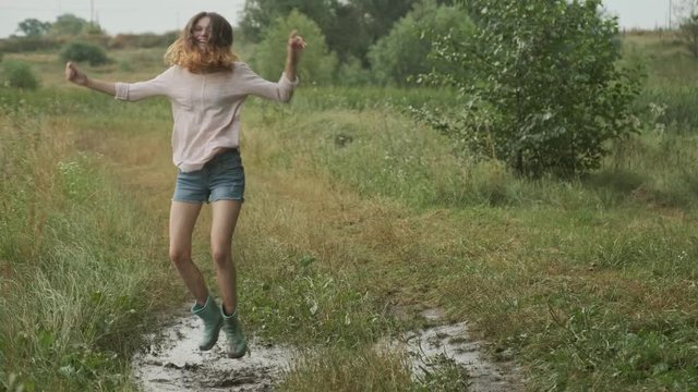 Beautiful smiling teen girl jumping in very muddy puddle on country road, summer nature background after rain