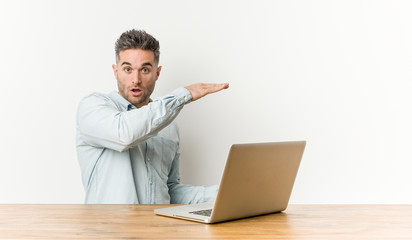 Young handsome man working with his laptop shocked and amazed holding a copy space between hands.