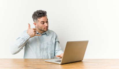 Young handsome man working with his laptop showing a mobile phone call gesture with fingers.