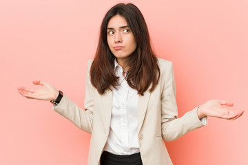 Young brunette business woman against a pink background doubting and shrugging shoulders in...