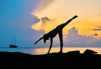 Silhouette of a young woman doing yoga exercises by the sea against the backdrop of colourful dawn	