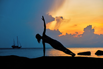 Silhouette of a young woman doing yoga exercises by the sea against the backdrop of colourful dawn	