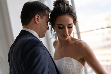 bride and groom hugging near window. feeling of love in newlyweds. guy and girl in wedding clothes pose in classical interior.