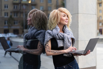 young hipster girl with tattoo on her arm and afro hair with laptop stands onstreet in break from...