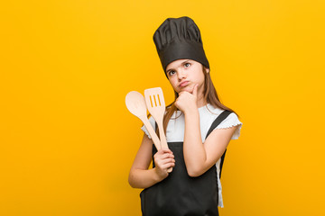 Little caucasian girl wearing a chef costume looking sideways with doubtful and skeptical expression.