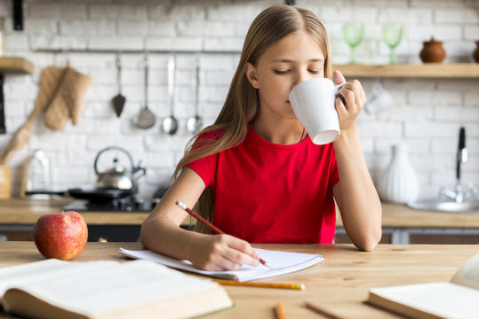 Schoolgirl Holding Cup Doing Homework At Kitchen Table