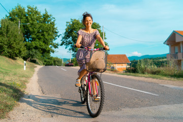 Young woman in a sunny summer day wearing dress riding a bike bicycle on the open road trough the village