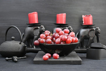 Still life: bunches of dark grapes in a bowl,grapes on a board,a piece,a teapot,a box,a candlestick in the form of three elephants with candles on a table with a black tablecloth on a black background