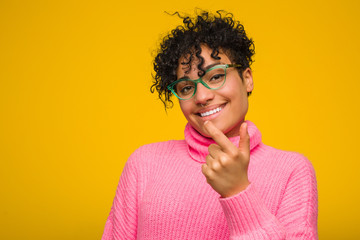 Young african american woman wearing a pink sweater pointing with finger at you as if inviting come closer.