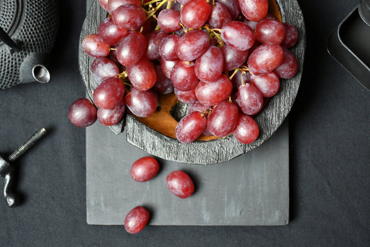 Top view: several bunches of dark grapes in a wooden bowl,three grapes nearby on a wooden board,a cast-iron piece,a teapot & a metal box on a table with a black tablecloth on a black wooden background