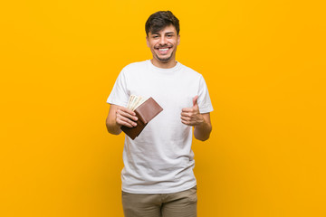 Young hispanic man holding a wallet smiling and raising thumb up