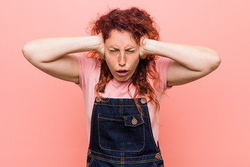 Young pretty ginger redhead woman wearing a jeans dungaree covering ears with hands trying not to hear too loud sound.