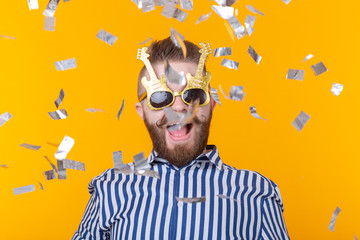 Joyful young hipster male in a paper cap and glasses is laughing happily among flying confetti on a yellow background. The concept of a mega party and holiday.
