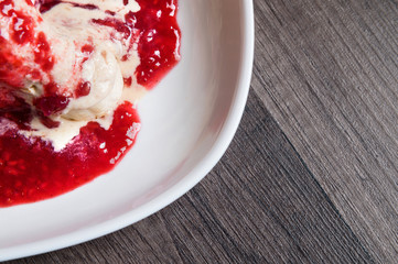 Closeup Vanilla ice cream poured with strawberry or raspberry jam with sliced banana slices on a white plate on a dark wooden table. You can do this at home on vacation