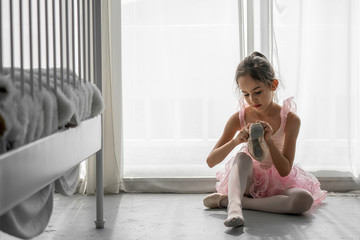 Cute beautiful young little girl ballerina dancer in pink leotard and tutu ballet dress costume sitting on the floor in bedroom wearing and tie ballet slipper shoe for private ballet dance practice. - obrazy, fototapety, plakaty