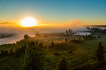 Orange sunny light with rays and shadows over the foggy river at dawn. The fog rises above water and stretches to the rising sun. Sunlit misty forest and river in nature
