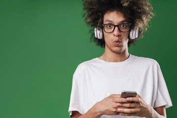 Photo closeup of perplexed man with afro hairstyle looking at camera while using cellphone and headphones