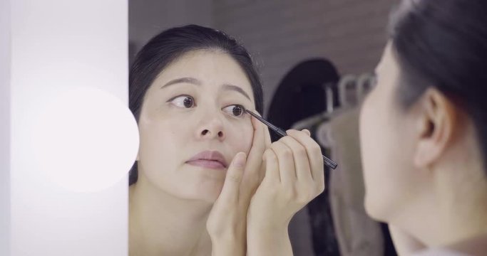 beautiful young dark haired asian woman entertainer using black eyeliner pen while applying makeup on face. close up reflection of girl model from dressing table mirror in backstage room before work