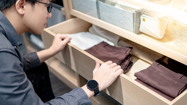 Asian Man Wearing Glasses Opening Closet Drawer Choosing Pants Or Trousers For Dressing Up. Home Lifestyle Concept