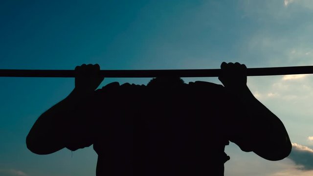 A young man makes pull-ups on the horizontal bar. Silhouette of a man against the background of dark blue cloudy sky.