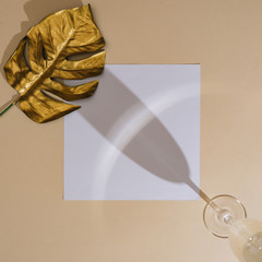 MInimal top view of champagne glass with golden monstera leaf and white paper card copy space. Summer creative concept.