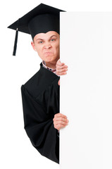 Angry male graduate student peeking from behind a blank panel, isolated on white background. Handsome upset graduate teen boy student in mantle showing empty placard board.