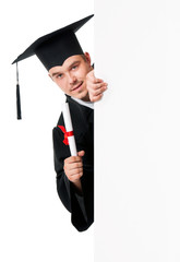 Male graduate student peeking from behind a blank panel, isolated on white background. Handsome graduate guy student in mantle showing blank placard board.