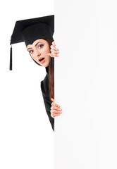 Surprised female graduate student peeking from behind a blank panel, isolated on white background. Beautiful graduate teen girl student in mantle showing blank placard board.