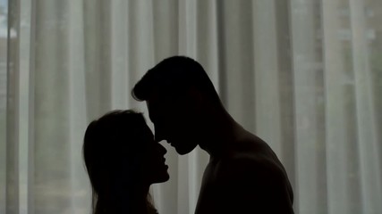 Close-up silhouette of a loving kissing couple on the background of the window. The man is shirtless and the girl in the white shirt standing in front of the window and hugging, silhouette.