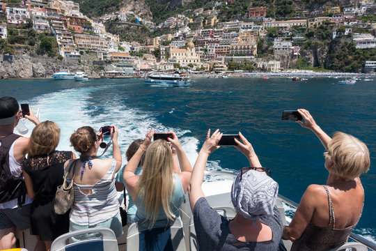 Tourists on a ferry boat as it leaves Positano take photographs on their smartphones of this Amalfi Coast town - Image