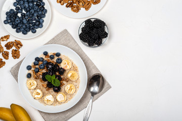 Oatmeal porridge with bananas, walnuts, blackberries, blueberries, honey and mint in a white bowl on a white background. Healthy breakfast and homemade diet food. Top view in flat lay style.