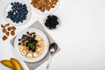 Oatmeal porridge with bananas, walnuts, blackberries, blueberries, honey and mint in a white bowl on a white background. Healthy breakfast and homemade diet food. Top view in flat lay style.