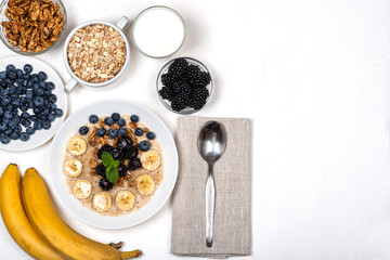 Oatmeal porridge with bananas, walnuts, blackberries, blueberries, honey and mint in a white bowl on a white background. Healthy breakfast and homemade diet food. Top view in flat lay style.