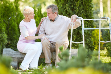 Full length portrait of senior man leaning on walker resting in park with caring nurse or wife,...