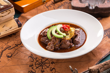 Braised beef served on white plate with chili pepper and avocado on old map background