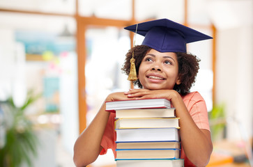 education, graduation and knowledge concept - happy smiling african american graduate student girl in bachelor cap with books dreaming over school background