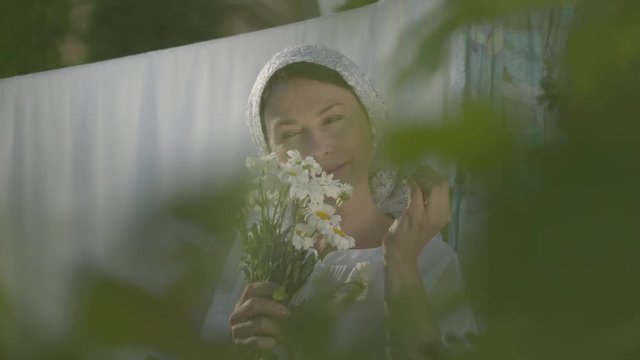 Attractive senior woman with white shawl on her head tears off daisy petals at the clothesline outdoors. Washday. Positive carefree housewife doing laundry. Shooting from behind tree brunch
