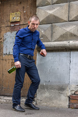 Drunk man with a serious look and with a bottle of alcohol is resting near the house after a working day