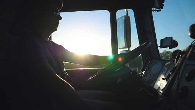 Silhouette of lorry driver driving through countryside in evening. Man in sunglasses controlling his truck attentive watching road. View inside cabin of lorry. Inside shot Slow motion Close up
