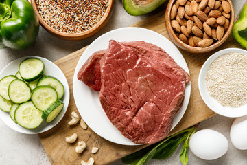 top view of raw meat on wooden chopping board near nuts, groats, eggs and green vegetables, ketogenic diet menu