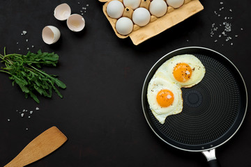 Fried eggs in frying pan with arugula leaves, salt and pepper for breakfast on black wooden...