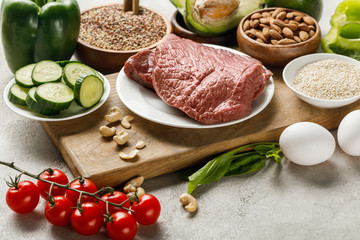 fresh raw meat on wooden chopping board near nuts, vegetables and groats, ketogenic diet menu