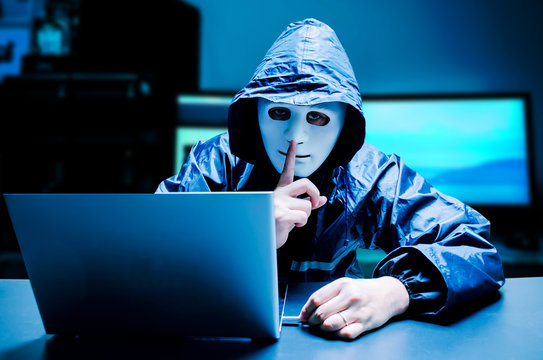 Anonymous computer hacker in white mask and hoodie. Obscured dark face making silence gesture try to hack and steal information system data from computer with usb thumb drive on dark background.